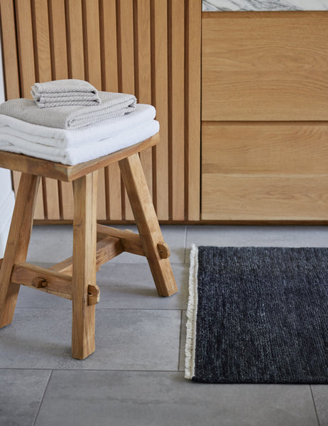 #color::white | The white turkish cotton Air Weight Towel Set by Coyuchi sits in a stack atop a wooden stool in a bathroom with wooden cabinetry and a gray rug