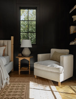 The Belmont taupe boucle accent chair sits next to a bed and a wooden side table atop a natural-toned pattered rug