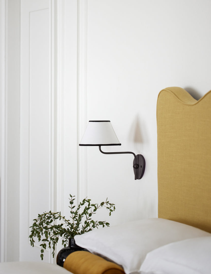 #color::black-trim #finish::bronze | The Magdalene black single sconce is mounted to the right of a golden linen bed on a white wall