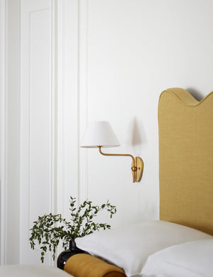 The Magdalene brass single sconce is mounted to the right of a golden linen bed on a white wall