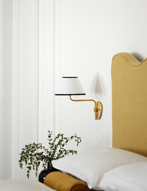 The Magdalene brass single sconce is mounted to the right of a golden linen bed on a white wall