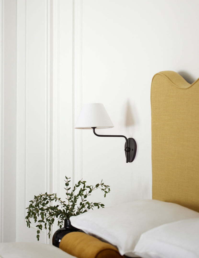 #color::white #finish::bronze | The Magdalene black single sconce is mounted to the right of a golden linen bed on a white wall