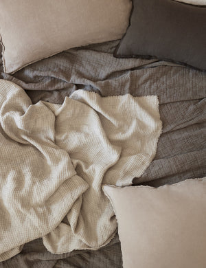 The Ojaj pebble cotton matelassé coverlet by pom pom at home lays amongst a gray throw blanket, a dark gray sham, and lighter gray throw pillows