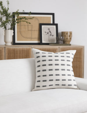 Bertu pumice gray pillow with a woven dash pattern by Bolé Road Textiles sits on a white sofa with a rattan cane sideboard in the background