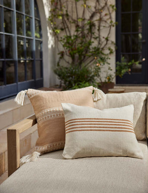 Marchesa sandstone indoor and outdoor square pillow with tasseled corners sits on a natural linen sofa with an orange striped throw pillow