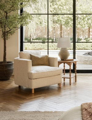 The Belmont taupe boucle accent chair sits atop chevron wooden floors next to a round wooden side table with sculptural legs