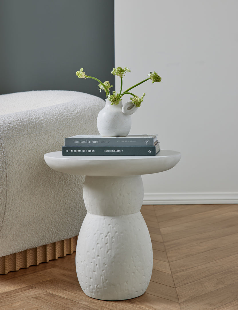 | The Clouded White Round Side Table sits in a studio with a white vase and stack of books next to a white boucle accent chair