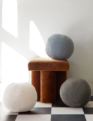 A blue, ivory, and khaki Bouclé Ball Pillow by Sarah Sherman Samuel sit on a black and white checkerboard floor and a cognac velvet ottoman