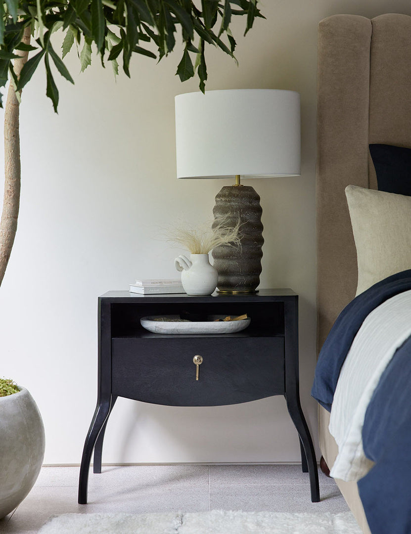 | The Anabella black wood nightstand has a lamp with a ribbed base, a stack of books, and a white vase sitting atop it