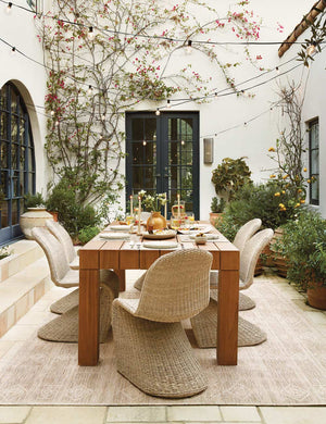 The Yamina taupe indoor and outdoor rug lays in an outdoor space under a rectangular dining table surrounded by wicker chairs