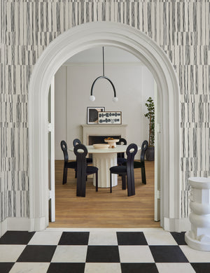 The Broken Stripe black and white Wallpaper by Sarah Sherman Samuel featuring offset organic stripes surrounds an entryway to a dining room featuring a white round pedestal dining table and sculptural black wooden dining chairs
