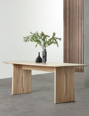 The Henrik light wood dining table sits in a studio atop a gray floor with two sculptural black vases sitting atop it