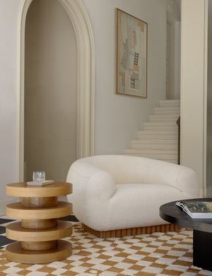 The Pentwater Four-Tiered Natural Round Side Table sits next to a boucle accent chair atop an ochre and white checkerboard rug