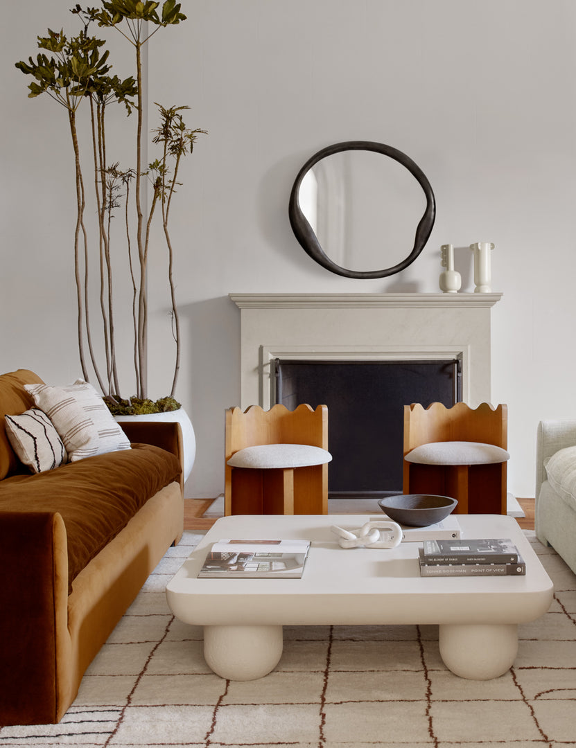 #color::black | The Doreen black organic round mirror is hung in a living room with a rounded sculptural coffee table, two sculptural accent chairs, and a cognac velvet sofa