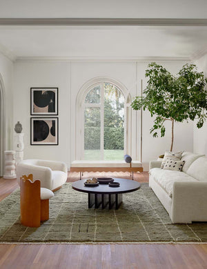 Pentwater black wooden Round Coffee Table sits atop an olive green patterned rug between a linen sofa and two accent chairs