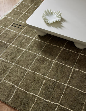 Khaki green Irregular Grid Rug by Sarah Sherman Samuel lays under a rounded coffee table with a shell-esque centerpiece bowl sitting atop it