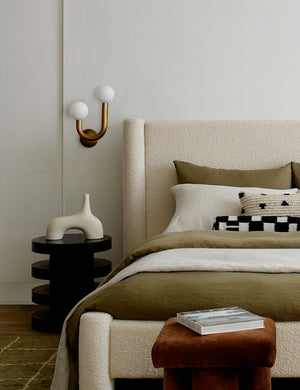 Natural-brass left-facing Happy Sconce by Regina Andrew is hung to the left of a boucle framed bed above a three tiered bed stand