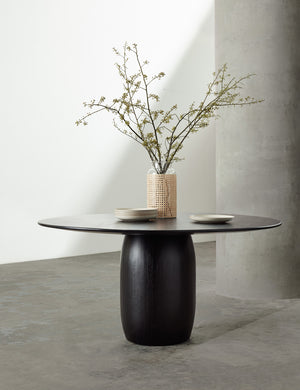 The Maroko black round dining table sits in a studio with a glass vase and a stack of plates
