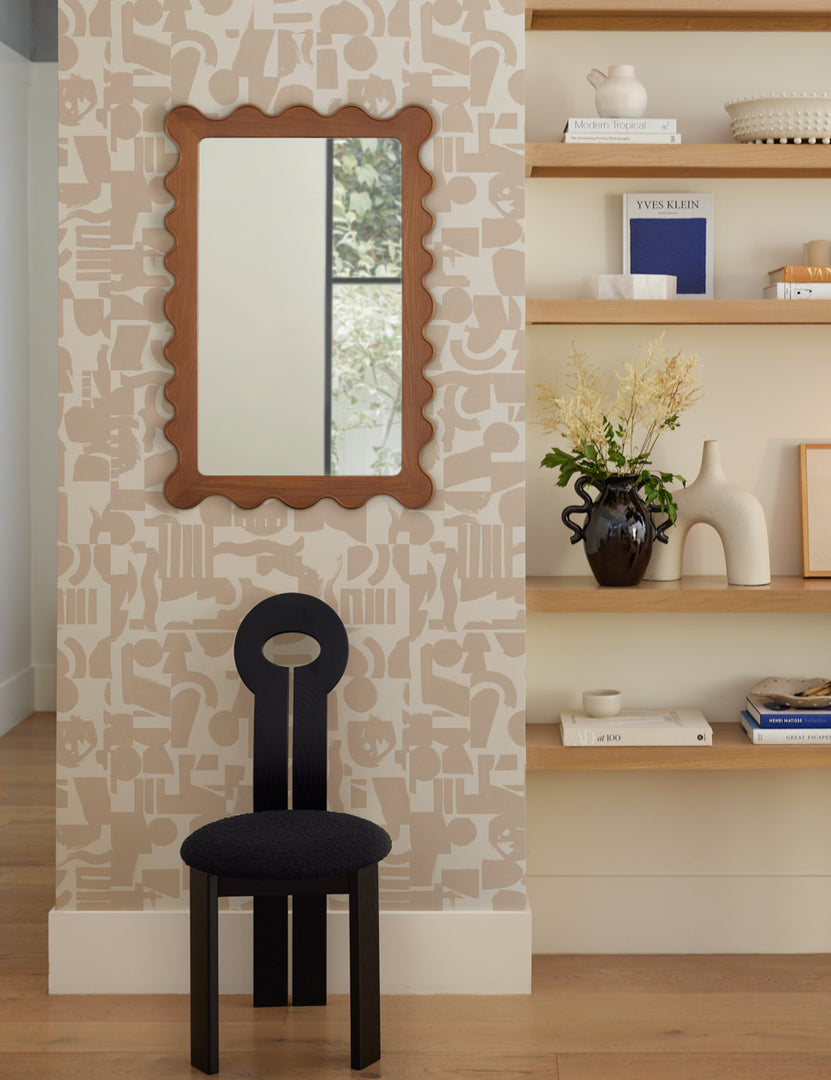 #color::honey-oak | The Ripple mirror hangs on a patterned wall above a black sculptural chair next to a shelf with decor