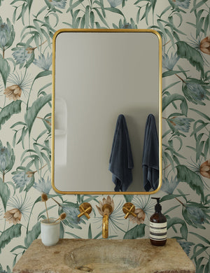 Green-toned Tropical Wallpaper by Rylee + Cru is in a bathroom with a rounded gold mirror and a stone sink with gold hardware