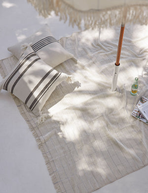 The Fez black and white indoor and outdoor throw pillow lays on an ivory outdoor blanket under a fringed beach umbrella