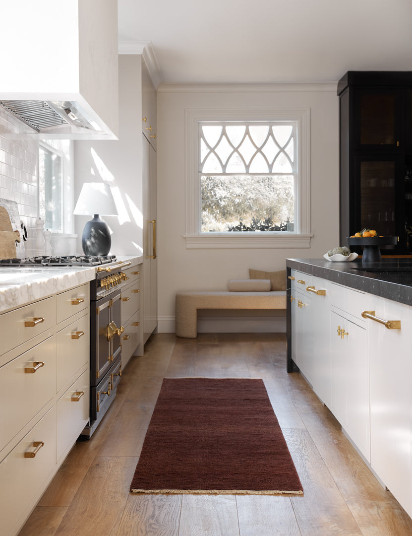 #color::brick #size::2-6--x-8- | The Heritage brick red runner rug lays in a kitchen with white cabinetry, gold hardware, and marble countertops 