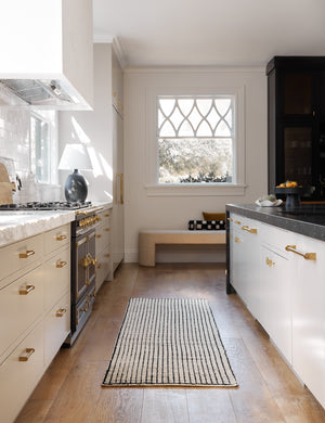 The Uma black and natural checkered grid pattern rug in its runner size lays in a kitchen with golden hardware and white cabinetry