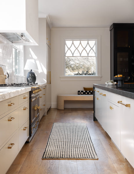 #color::black-and-natural #size::2-6--x-8- | The Uma black and natural checkered grid pattern rug in its runner size lays in a kitchen with golden hardware and white cabinetry