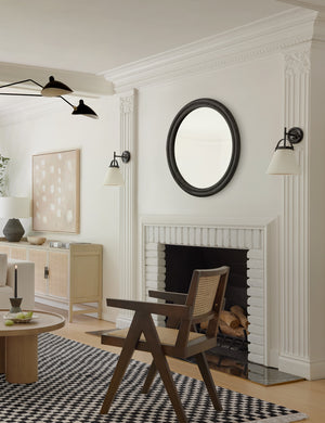 The Bourdon Double-Framed Black Round Mirror hangs on a white wall above a fireplace and a checkerboard rug