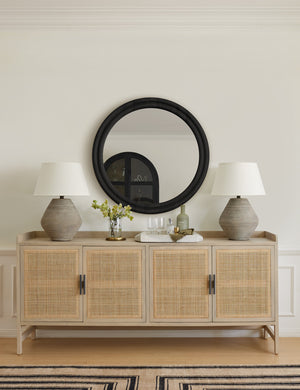 The Bourdon Double-Framed Black Round Mirror hangs on a cream wall above a rattan sideboard with lamps and decorative objects