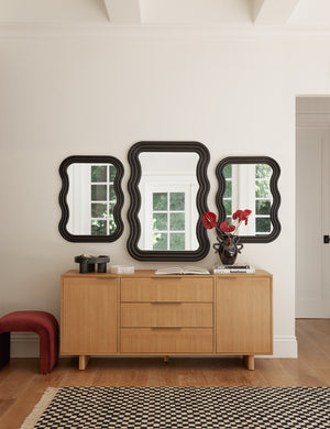 Three Wendolyn wavy thick-framed black wall mirrors hang above a light wood dresser and a black and white checkerboard rug.