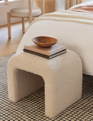 The Tate Cream Boucle stool sits at the end of a bed on a gridded rug with a wooden bowl and a stack of books sitting atop it