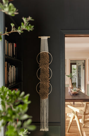 The Studio Nom Lwazi light brown woven Wall Hanging by nom hangs in a black painted room next to a bookshelf and a dining room in the background