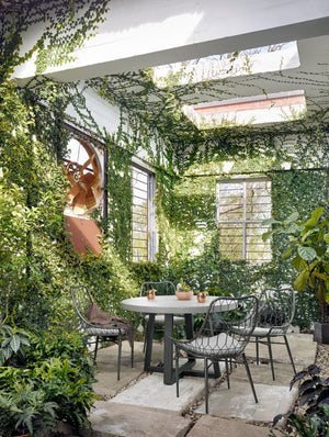 The Agatha indoor and outdoor dining table with lavastone top and black iron base sits in a covered outdoor space with ivy-covered walls and black woven dining chairs