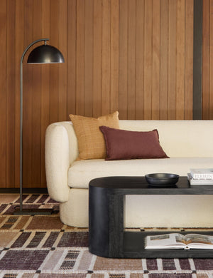 The Otto oil rubbed bronze arched floor lamp by Regina Andrew sits in a retro living room with wood paneled walls, a warm red-toned area rug, and a white boucle rounded sofa