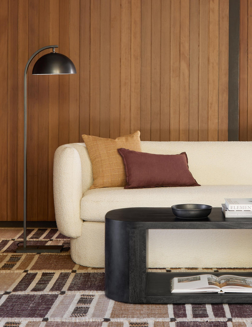 #color::oil-rubbed-bronze | The Otto oil rubbed bronze arched floor lamp by Regina Andrew sits in a retro living room with wood paneled walls, a warm red-toned area rug, and a white boucle rounded sofa