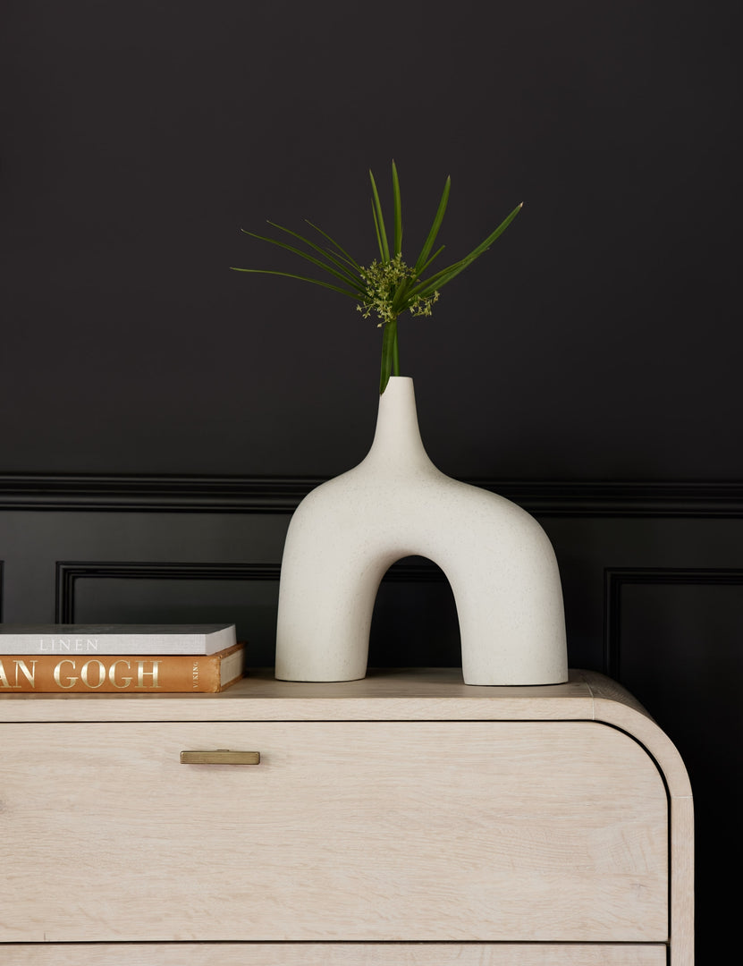 #color::white #size::large | The Leonor larger sculptural arched matte white ceramic Vase sitting atop a white washed dresser next to a stack of books in front of a black accented wall