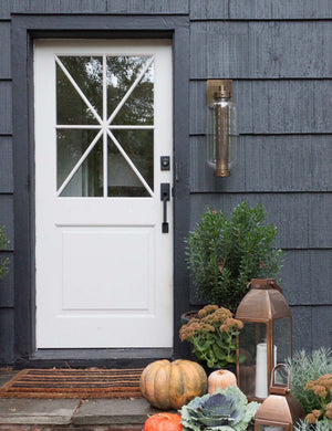 The Romelia indoor and outdoor sconce with metal mesh encased bulb and brass hardware hangs out the exterior of a black paneled home to the right of a white door