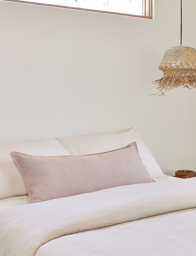 #color::greige #style::extra-long #fill type::with-feather-down-insert #fill type::pillow-cover-only | The arlo Greige long lumbar pillow lays on a bed with ivory linens with a jute pendant light hanging next to it