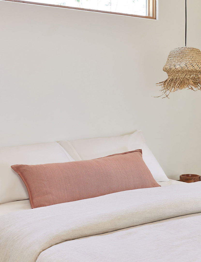 #color::terracotta #style::extra-long #fill type::with-feather-down-insert #fill type::pillow-cover-only | The arlo Terracotta long lumbar pillow lays on a bed with ivory linens with a jute pendant light hanging next to it