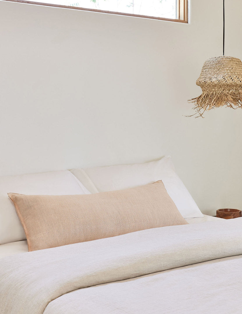 #color::blush #style::extra-long #fill type::with-feather-down-insert #fill type::pillow-cover-only | The arlo Blush pink long lumbar pillow lays on a bed with ivory linens with a jute pendant light hanging next to it