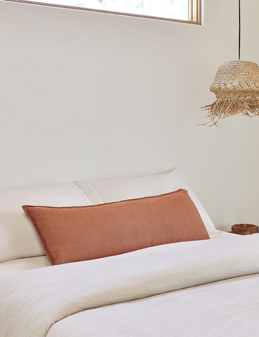 #color::rust #style::extra-long #fill type::with-feather-down-insert #fill type::pillow-cover-only | The arlo rust orange long lumbar pillow lays on a bed with ivory linens with a jute pendant light hanging next to it