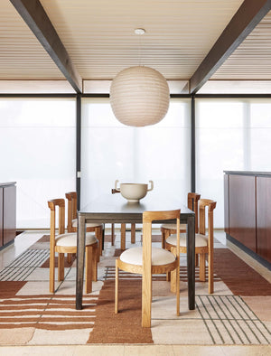 The Benita natural toned hand-knotted floor rug by Nina Freudenberger lays in a dining room with a black wooden dining table, sculptural dining chairs, and a white centerpiece bowl