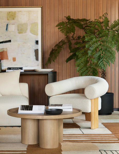 | The Delta ash wood round coffee table with three column legs sits in a living room with wood paneled walls, a brown geometric patterned rug and a rounded sherpa accent chair.