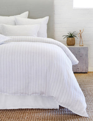 The Blake white and ocean linen duvet by pom pom at Home lays atop a white linen framed bed in a bedroom with a jute rug and a cubical gray nightstand