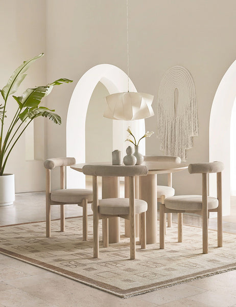 #color::whitewash | Mojave Round Dining Table sits in a cream toned room surrounded by cream cushioned dining chairs atop a cream and neutral patterned rug