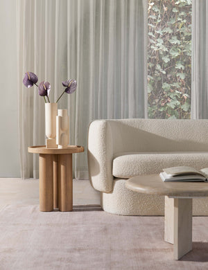 The Delta natural wooden side table with pedestal base sits in a bright living room with a rounded white boucle couch, an ivory plush rug, and a whitewashed oval coffee table