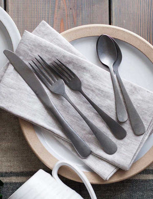 The Woodstock stainless steel 5-piece flatware set by farmhouse pottery sits on a dining table atop a ceramic plate and beige linen napkins