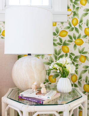 The Vivienne tan ceramic table lamp with spherical base sits atop a glass and woven side table with a white vase, a stack of books, and lemon wallpaper