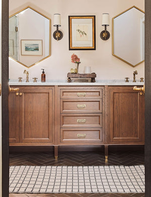 The Sebou natural and black dotted machine washable mat in its runner size lays in a bathroom with hexagonal golden framed mirrors